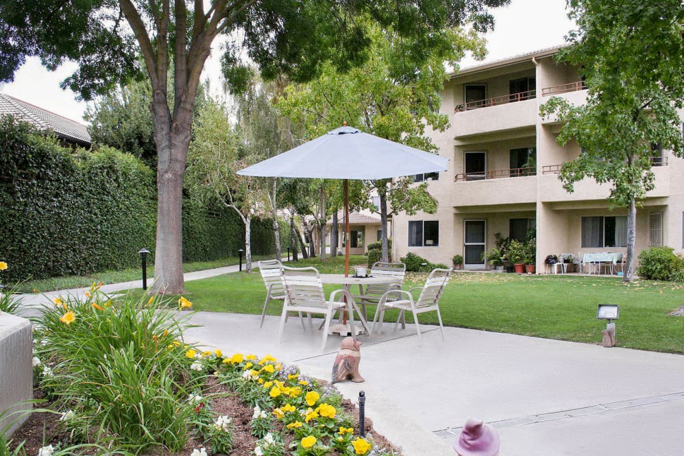 A serene outdoor patio area with tables and chairs under an umbrella, surrounded by flowers and trees, in front of a multi-story building.