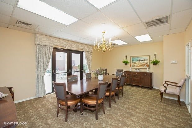 A well-lit dining room with a large conference table and eight chairs, featuring a chandelier, curtains, artwork on the wall, and a sideboard.