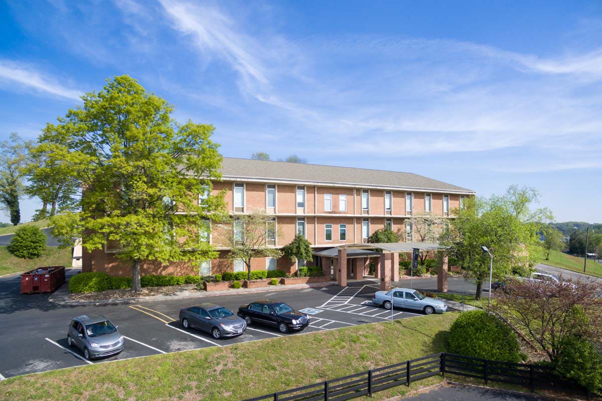 A hotel with a parking lot and surrounding trees under a clear blue sky.