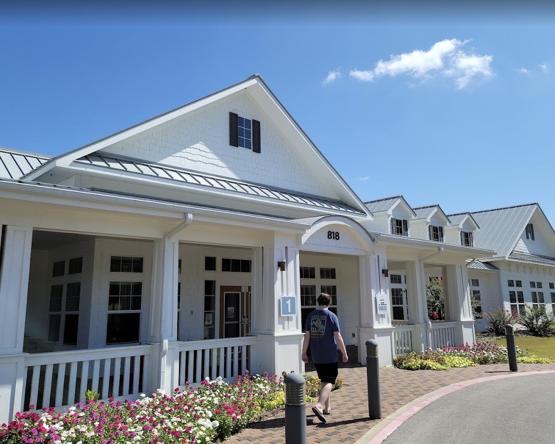 A person walking towards a white building with a porch on a sunny day.