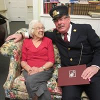 An elderly woman sits on a couch with a uniformed firefighter, both smiling warmly.