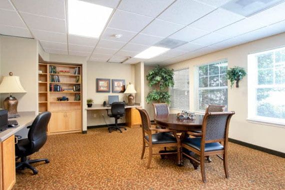A cozy office meeting room with a round table, chairs, bookshelf, and large windows.