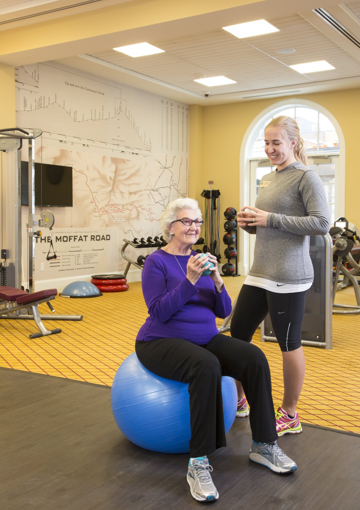 An elderly woman sitting on a balance ball in a gym, accompanied by a younger woman in workout attire.