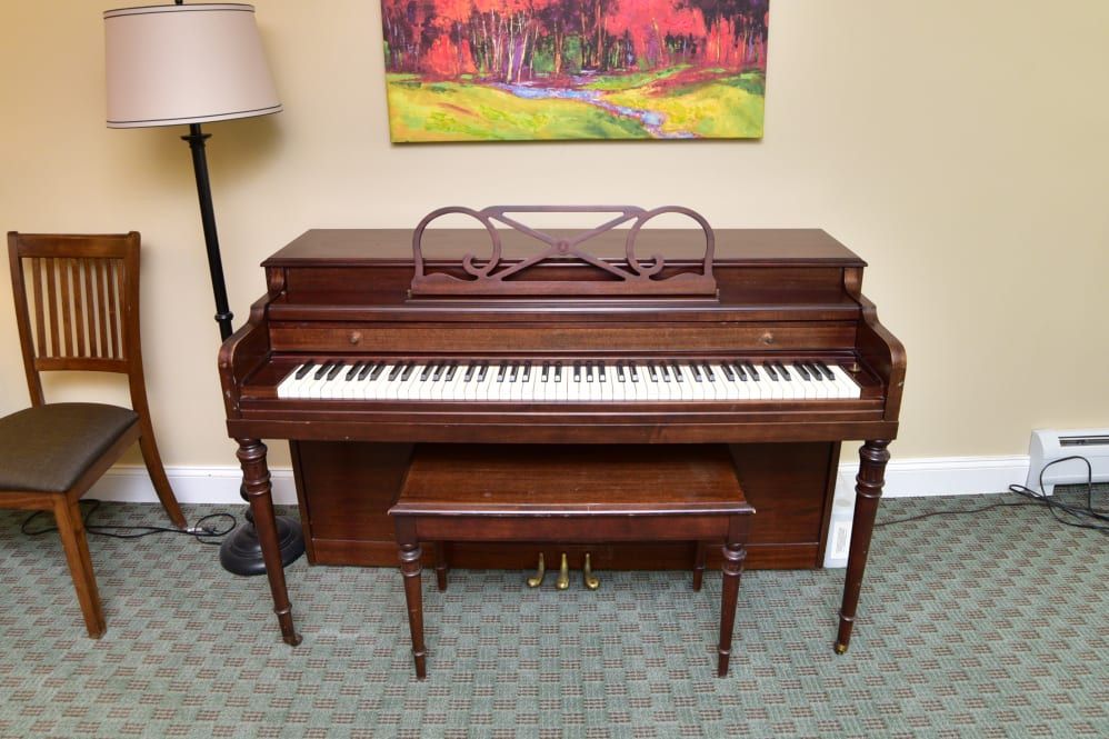 A wooden upright piano with a matching bench, positioned in a room with a floor lamp and a colorful painting on the wall.