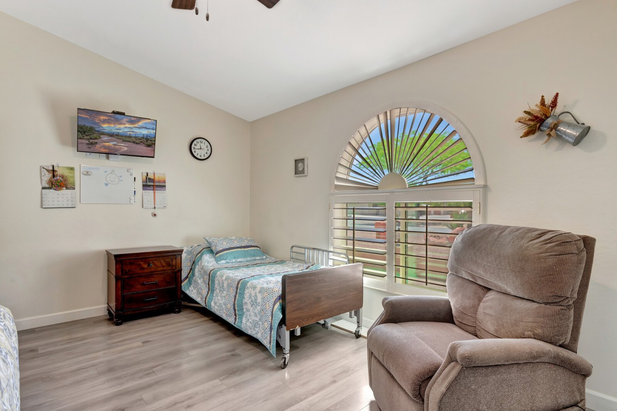 A cozy bedroom featuring a single bed, comfortable chair, wooden flooring, and a large window with sunlight streaming in.