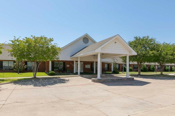 A single-story building with a prominent entrance and surrounding trees, captured on a sunny day.