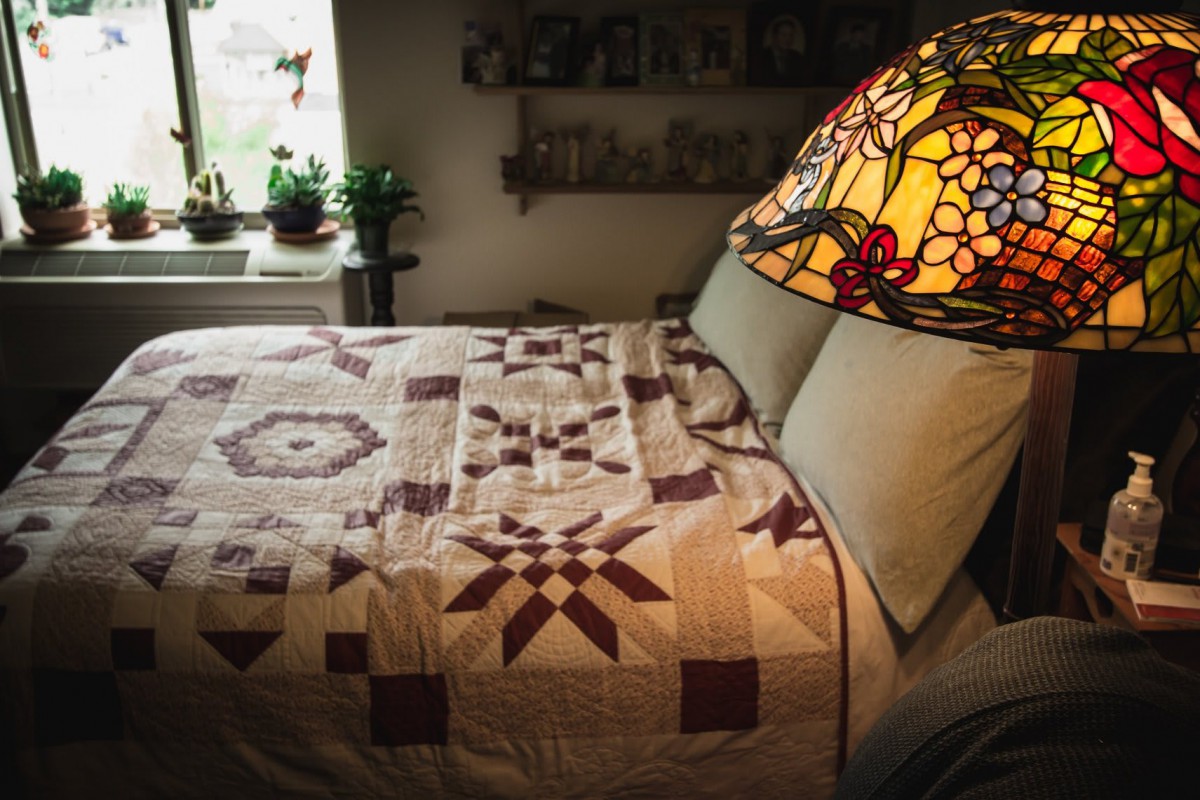 A cozy bedroom with a patterned quilt, stained glass lamp, and potted plants on the windowsill.
