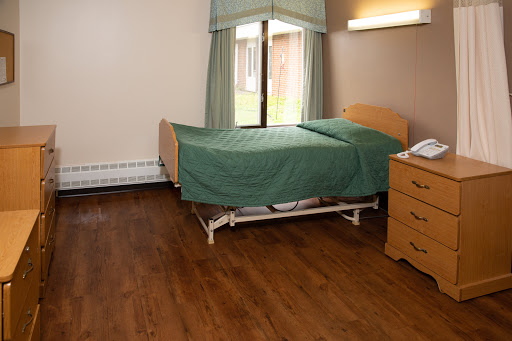 A hospital patient room featuring a bed with green bedding, wooden flooring, and natural light coming in through the window.