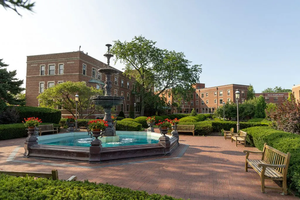 A peaceful park scene with a central fountain, surrounded by benches and greenery, set against the backdrop of brick buildings on a sunny day.