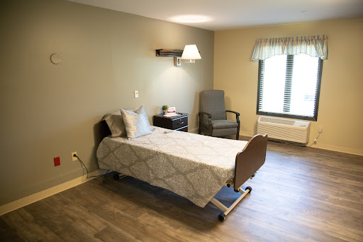 A clean and simple hospital patient room featuring a bed, a chair, and natural lighting.