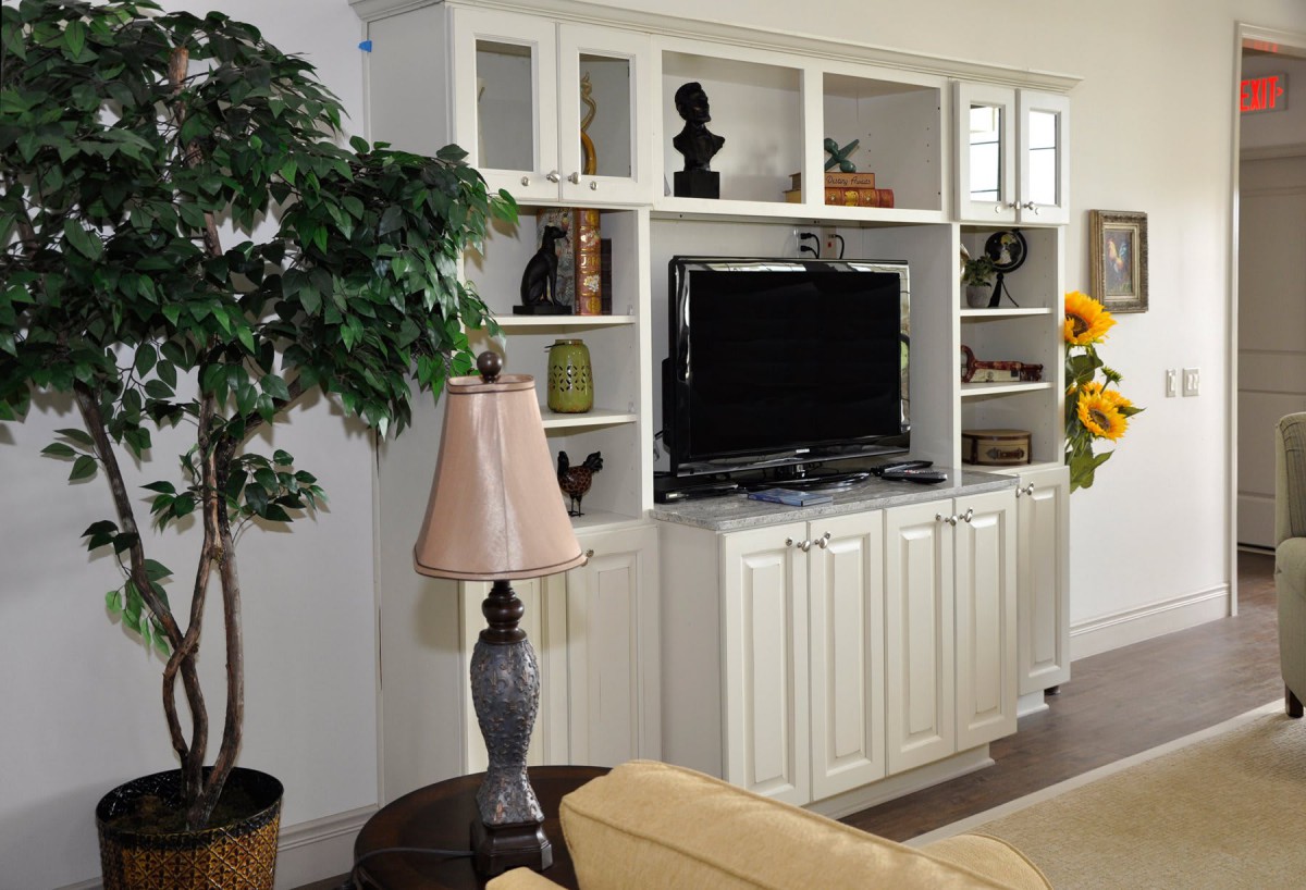 A cozy living room with a white entertainment center, featuring a television, decorative items on shelves, and a nearby table lamp.