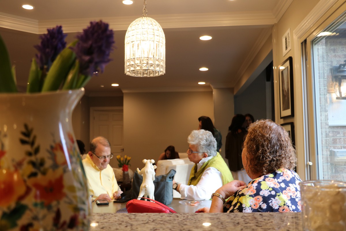 A group of people gathered around a kitchen table, engaged in conversation under a chandelier.