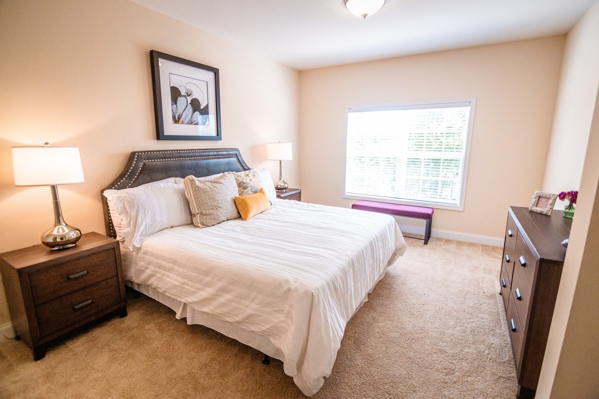 A cozy bedroom with a neatly made bed, soft carpet, and large window allowing natural light.