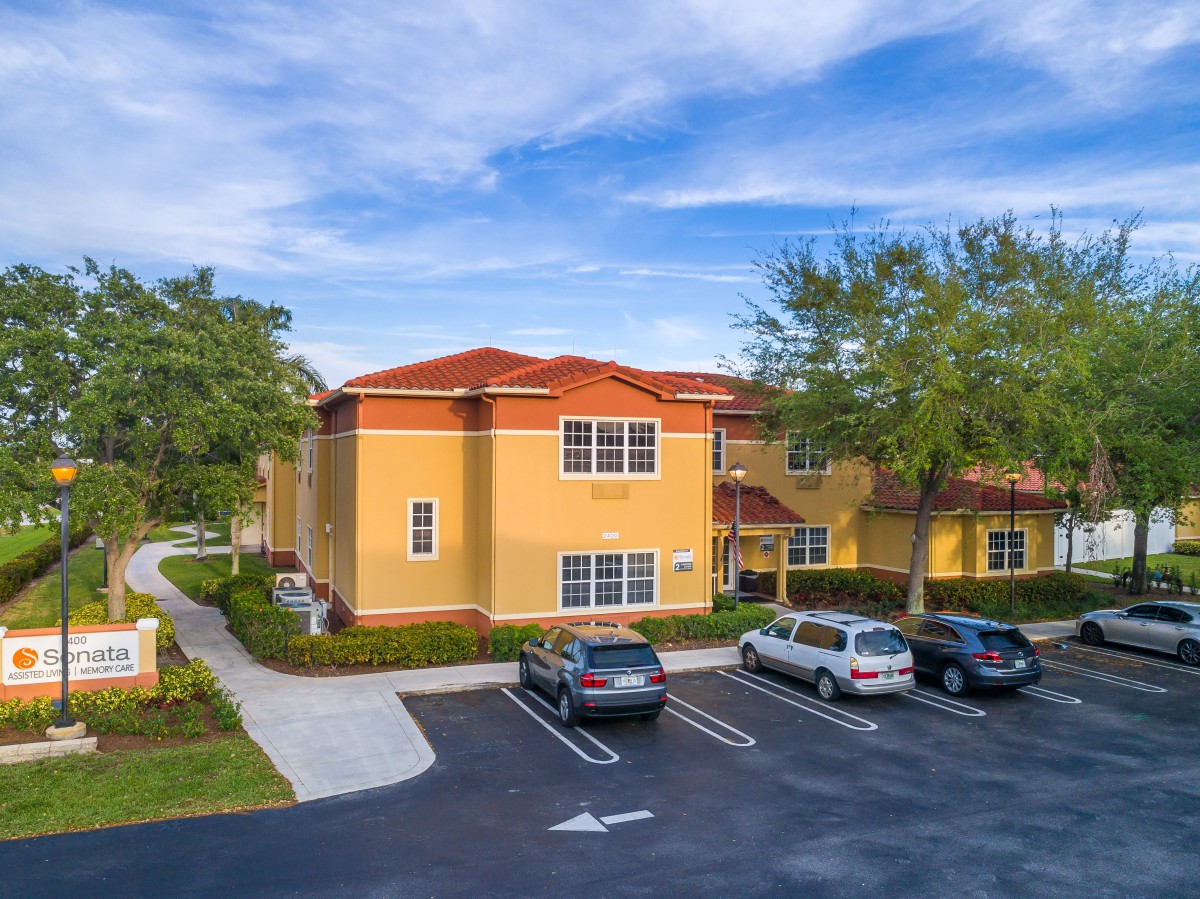 A suburban apartment complex with a parking lot and surrounding greenery under a partly cloudy sky.