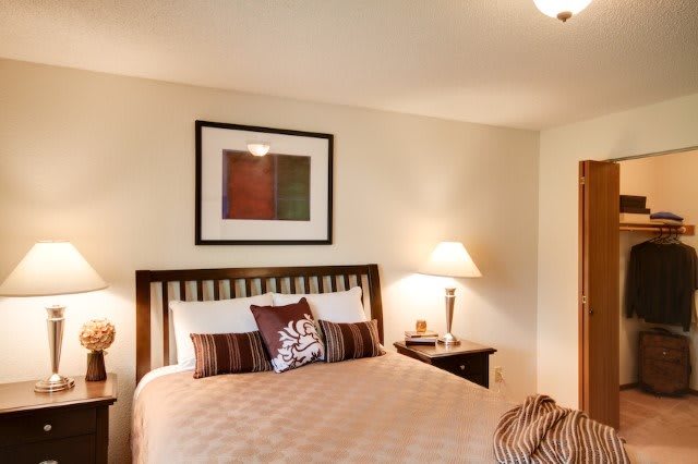 A cozy bedroom with modern decor, featuring a bed with decorative pillows, a nightstand with a lamp, and wall art.