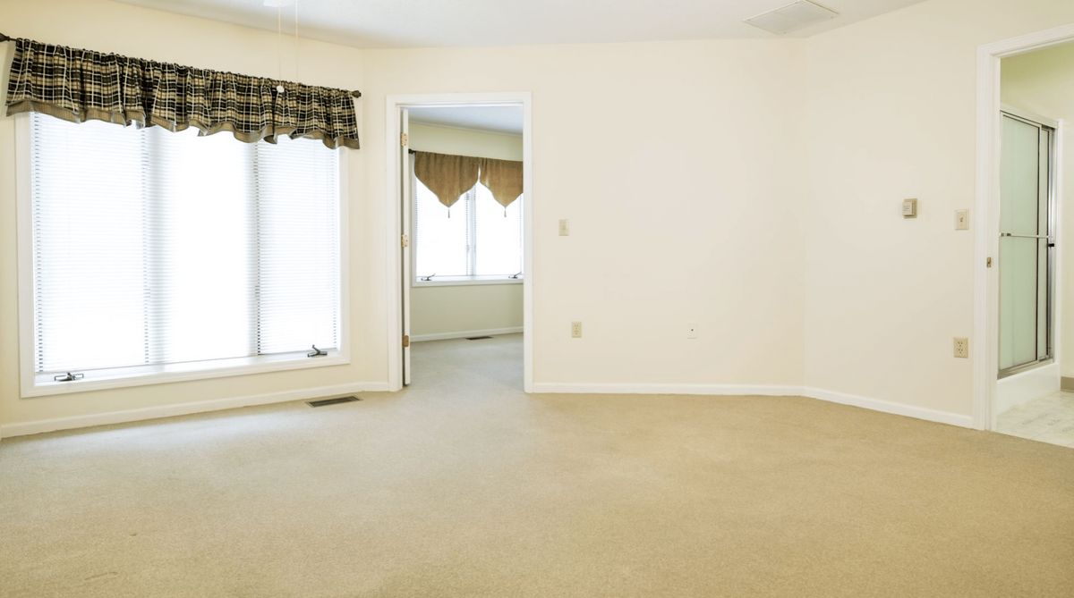 An empty room with carpet flooring and large windows covered by curtains.
