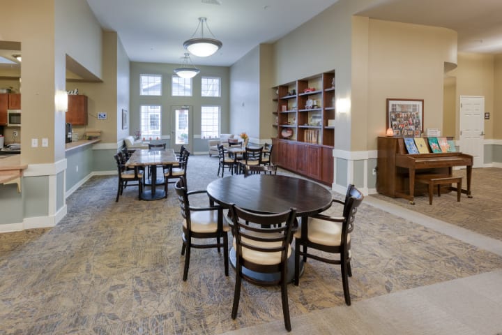 A well-furnished library room with tables and chairs, featuring bookshelves and ample natural light.