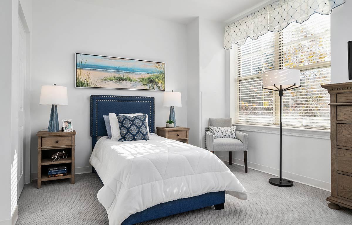A modern bedroom with a single bed featuring a blue headboard, complemented by a white duvet. The room is decorated with matching nightstands, an armchair, and a floor lamp. A landscape painting adorns the wall above the bed.