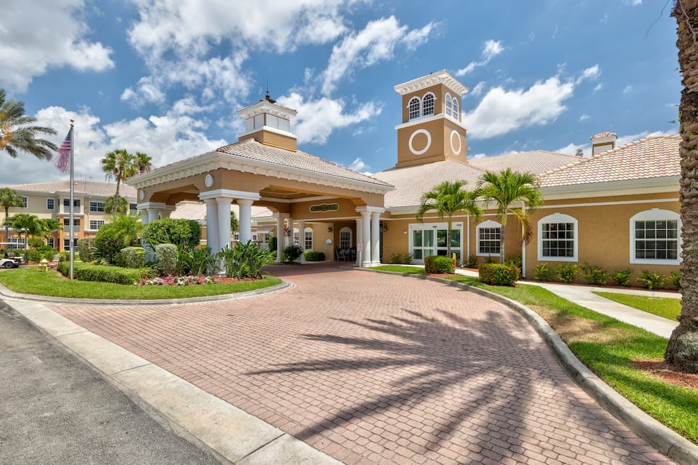 Elegant building entrance with palm trees and a cobblestone driveway under a partly cloudy sky.