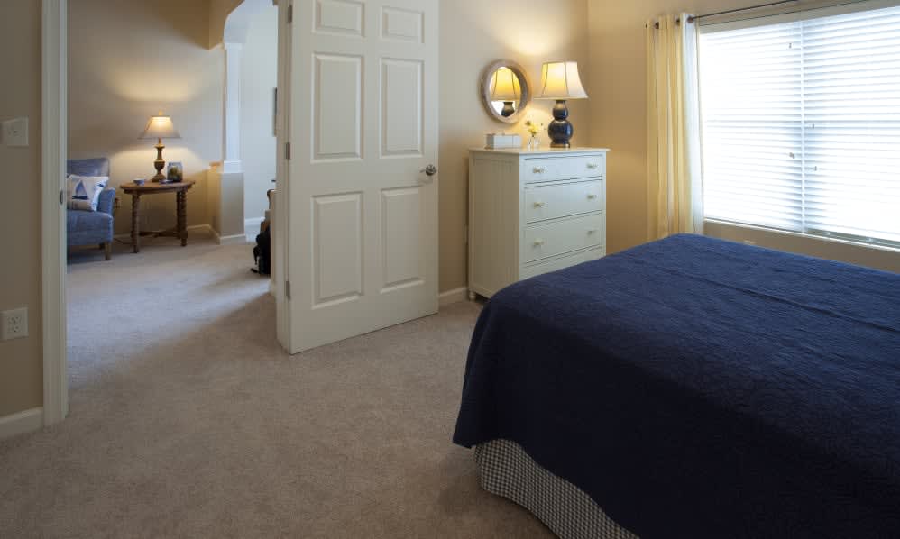 A cozy bedroom with a neatly made bed, a dresser with a lamp on top, and carpeted flooring.