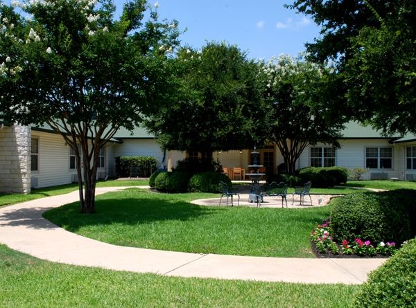 A peaceful garden scene featuring a curving pathway, lush greenery, and an outdoor seating area surrounded by trees.