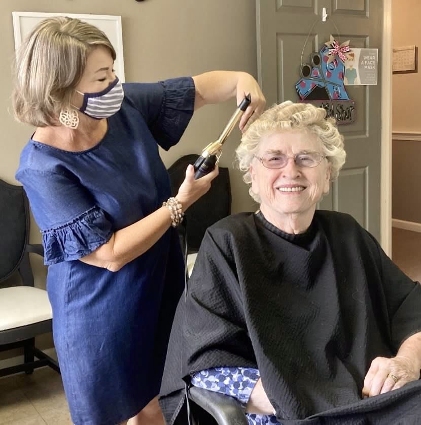 A hairstylist giving a haircut to a senior woman who is smiling, while wearing protective masks in a hair salon.