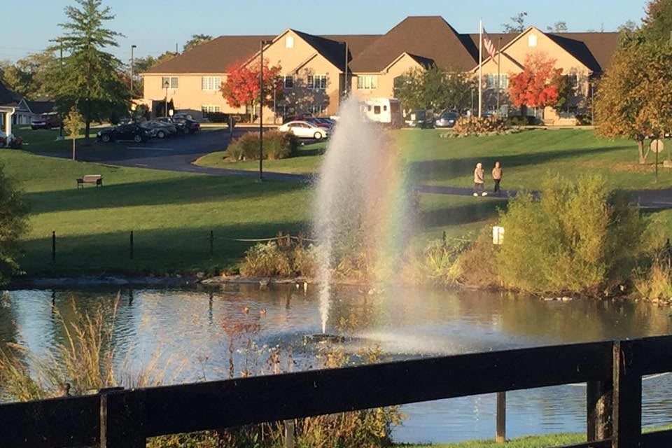 A serene park scene with a fountain creating a rainbow effect over a lake, surrounded by suburban homes and autumn trees.