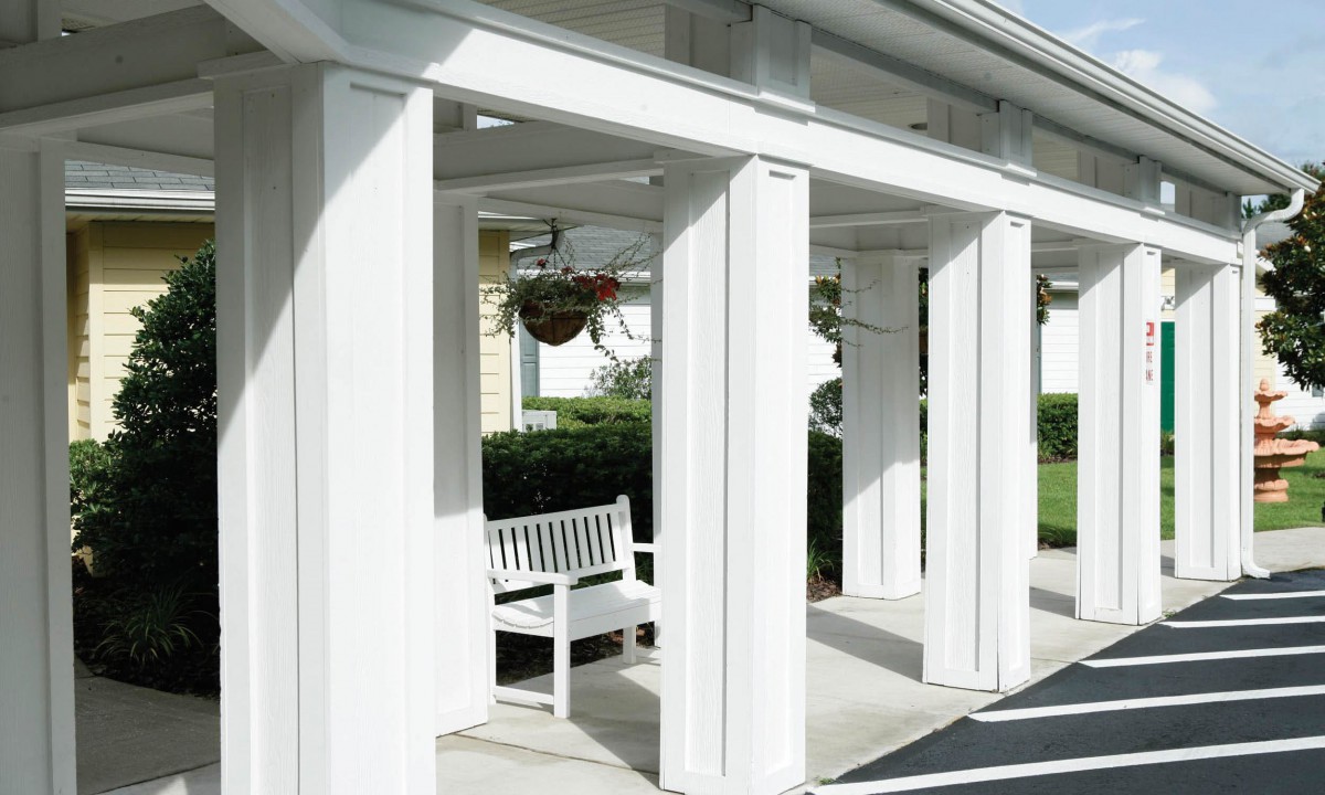 A white outdoor structure with columns, a bench, and hanging plants on a sunny day.