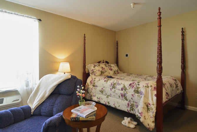 A cozy bedroom with a floral-patterned bed, a comfortable armchair, and a small side table with a lamp.