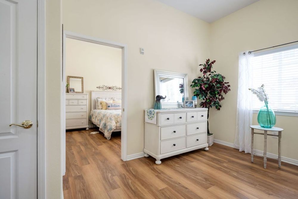 A spacious bedroom with a dresser and a large mirror, featuring wood flooring and bright lighting.