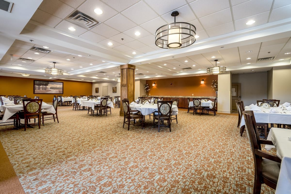 An elegant dining area with round tables and chairs, featuring warm lighting and carpeted flooring.