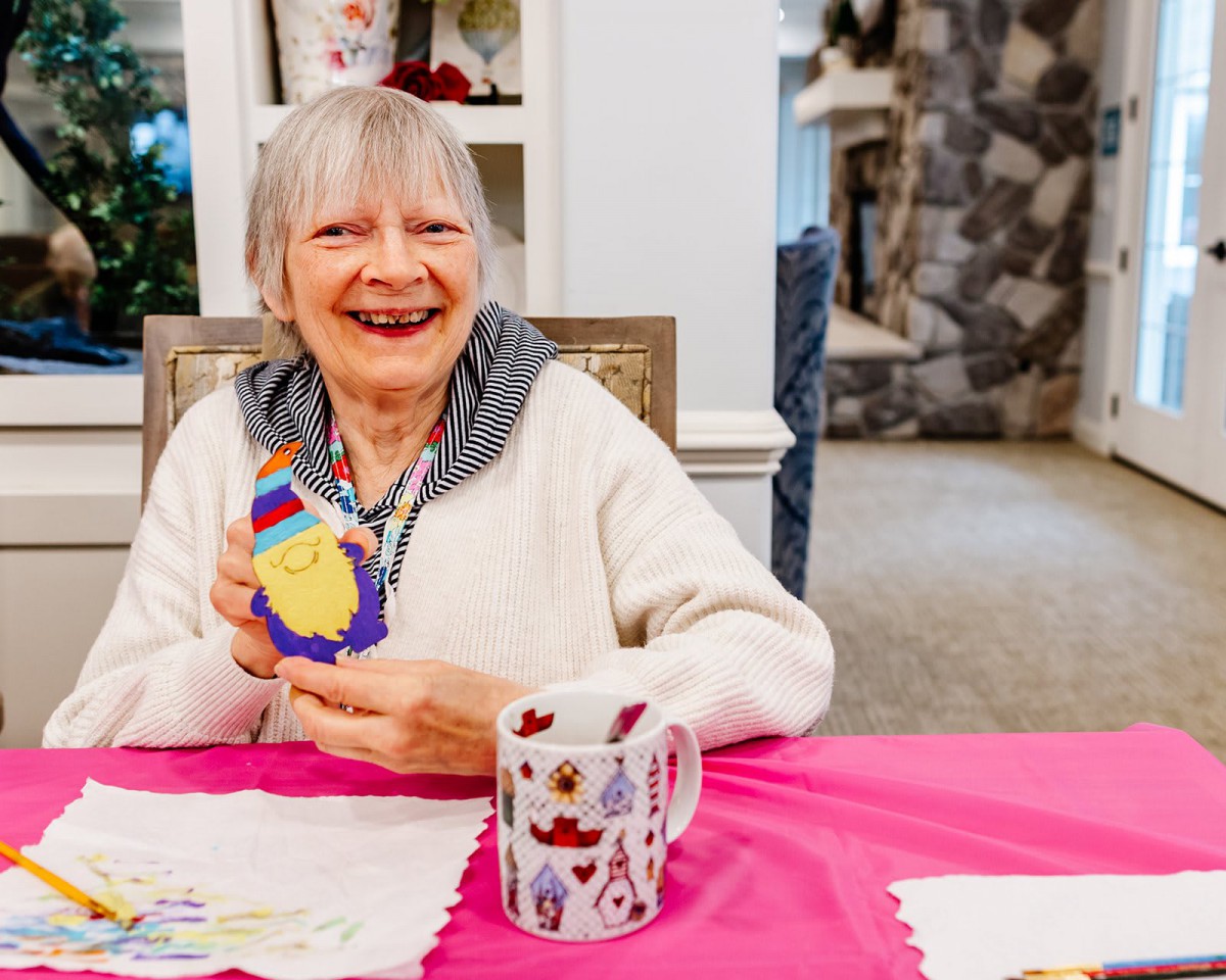 An elderly woman happily crafting at a table with a colorful mug.