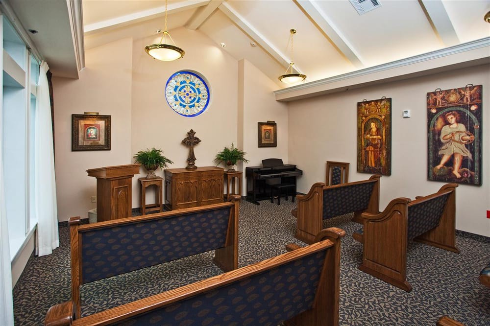 A small chapel interior with pews, an altar, and a piano, featuring a stained glass window.