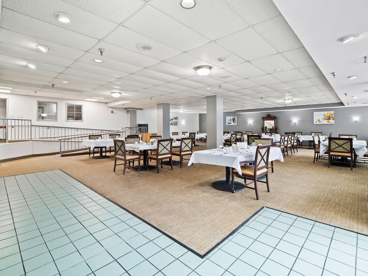 A spacious indoor dining area with several tables and chairs, featuring white tablecloths and a mix of carpet and tile flooring.