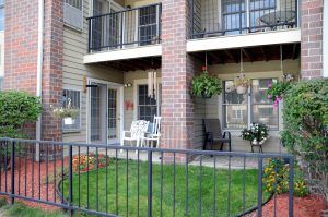 A cozy apartment balcony with outdoor furniture, hanging and potted plants, bordered by a black fence and a small green lawn.