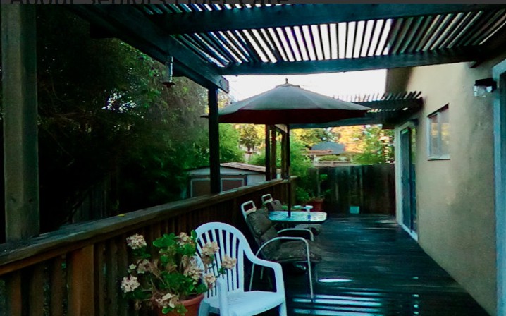 A cozy backyard patio with seating under a pergola, featuring chairs, a table, and vibrant plants.