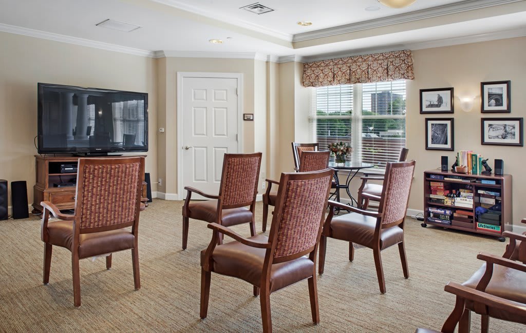 A meeting room with several chairs arranged in a semi-circle facing a television, featuring large windows that let in natural light.