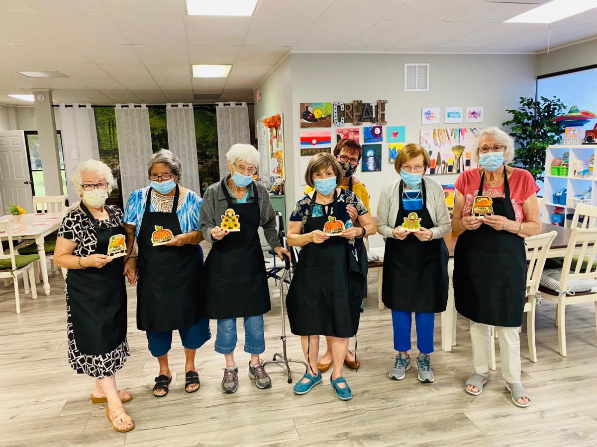 A group of elderly women in a cooking class, wearing aprons and masks.