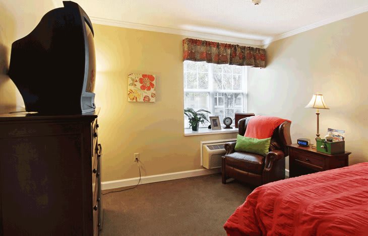 A cozy bedroom with a television, comfortable chair, and a window letting in natural light.