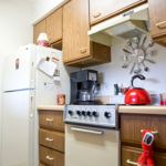 A retro-style kitchen featuring wooden cabinets, a stove, refrigerator, and small appliances like a coffee maker and red kettle.