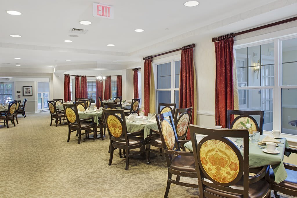 An elegant dining room with multiple tables and chairs, featuring warm lighting and red curtains.