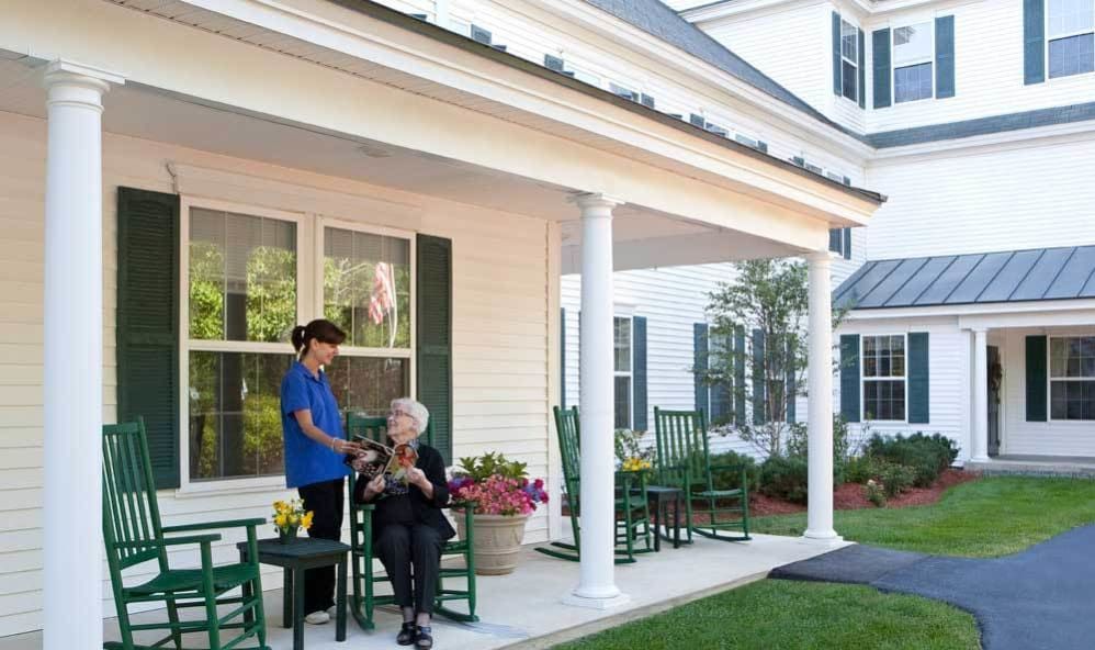 A caregiver and a senior person talking on a house porch surrounded by greenery.