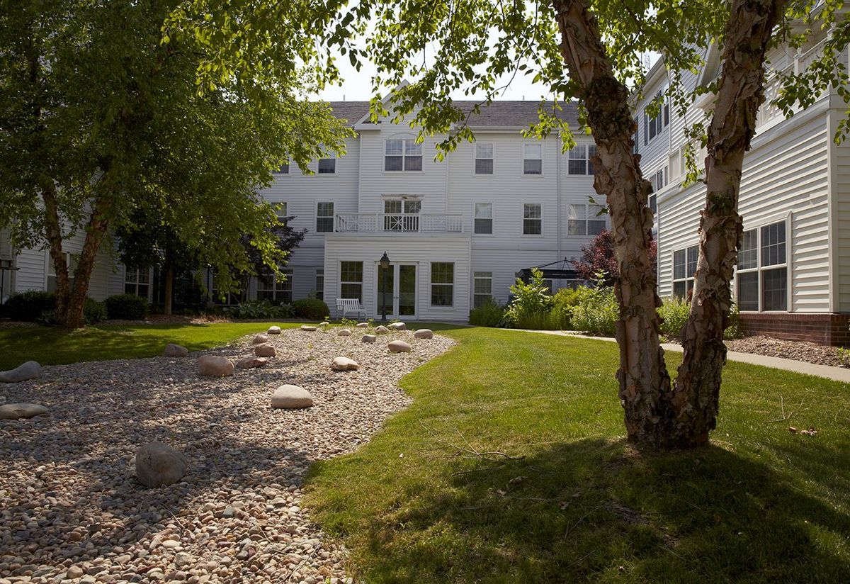 A serene residential garden with trees and a stone pathway leading to a large white building.