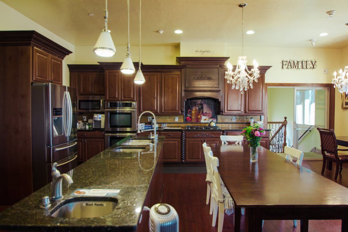 A modern kitchen with wooden cabinetry and granite countertops, featuring a large island and dining table under elegant lighting fixtures.