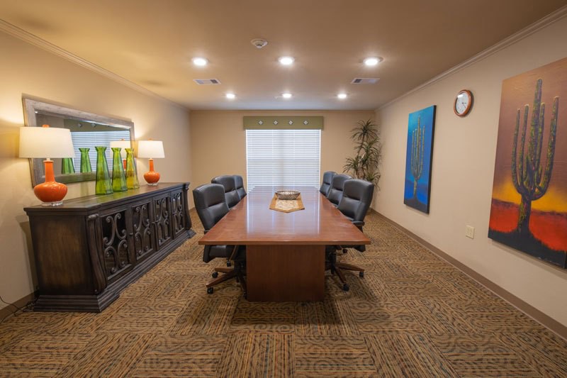 A well-lit conference room with a central wooden table surrounded by chairs, artwork on the walls, and window blinds.
