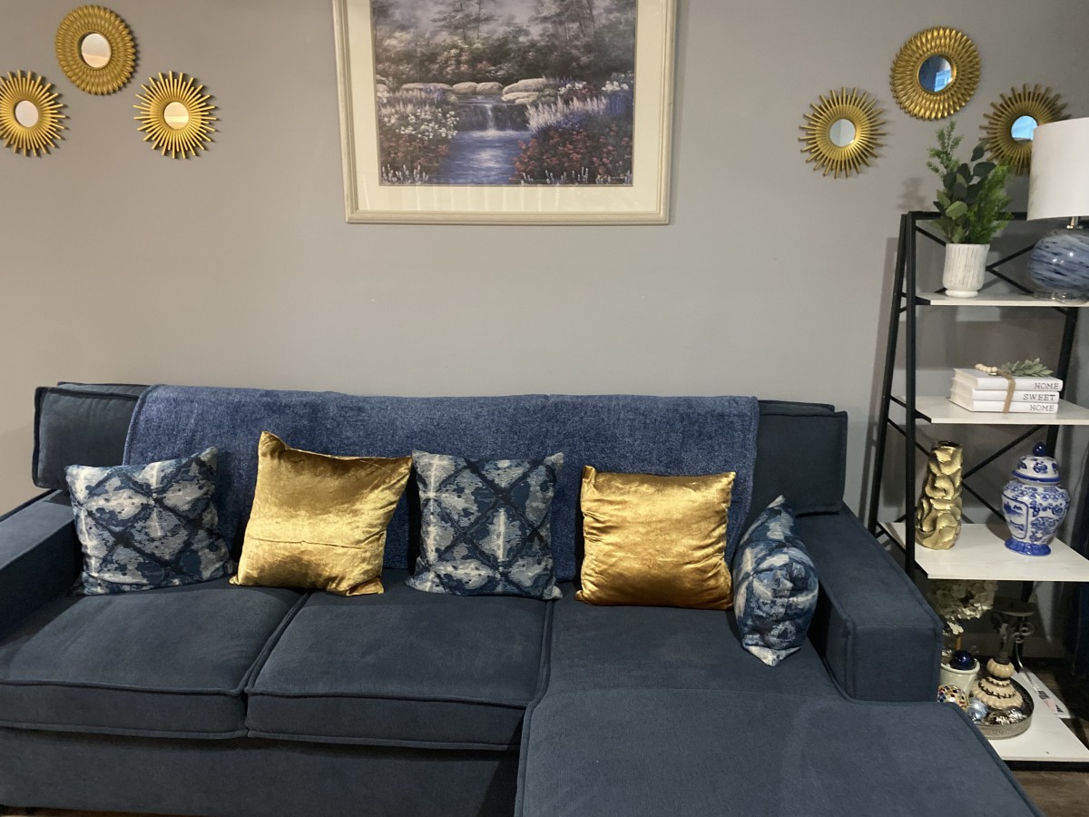 A cozy living room with a blue sofa adorned with golden and patterned cushions, accompanied by wall art and decorative items on a shelf.
