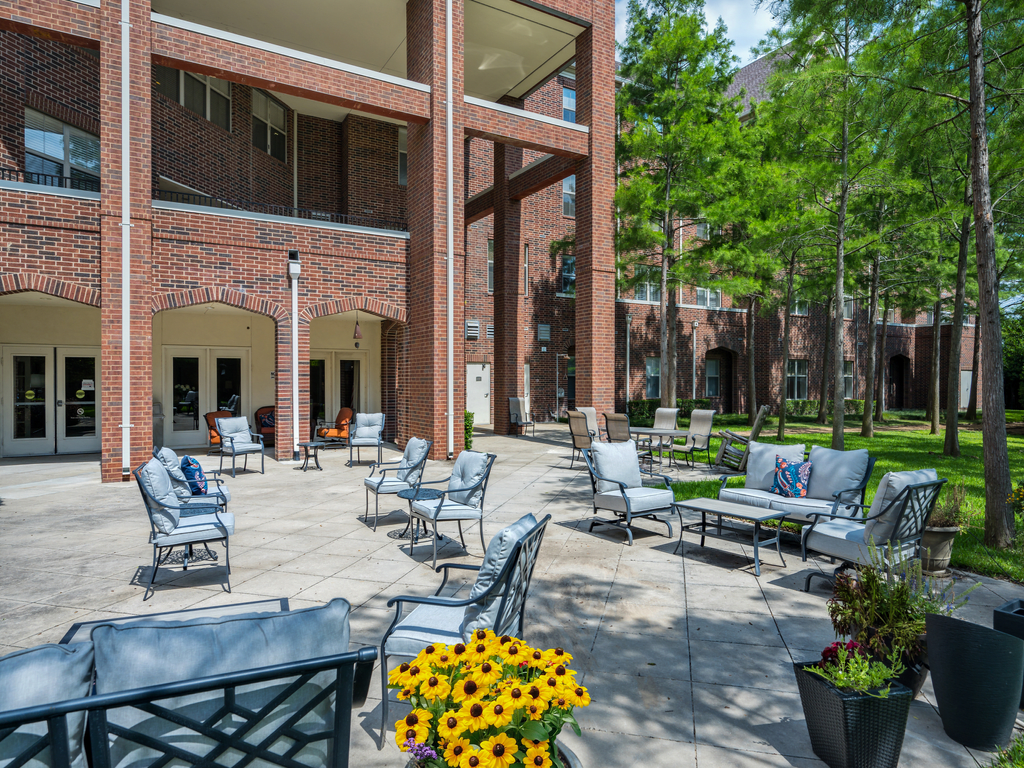 An inviting outdoor patio area with comfortable seating, surrounded by sunflowers and trees, adjacent to a brick building.