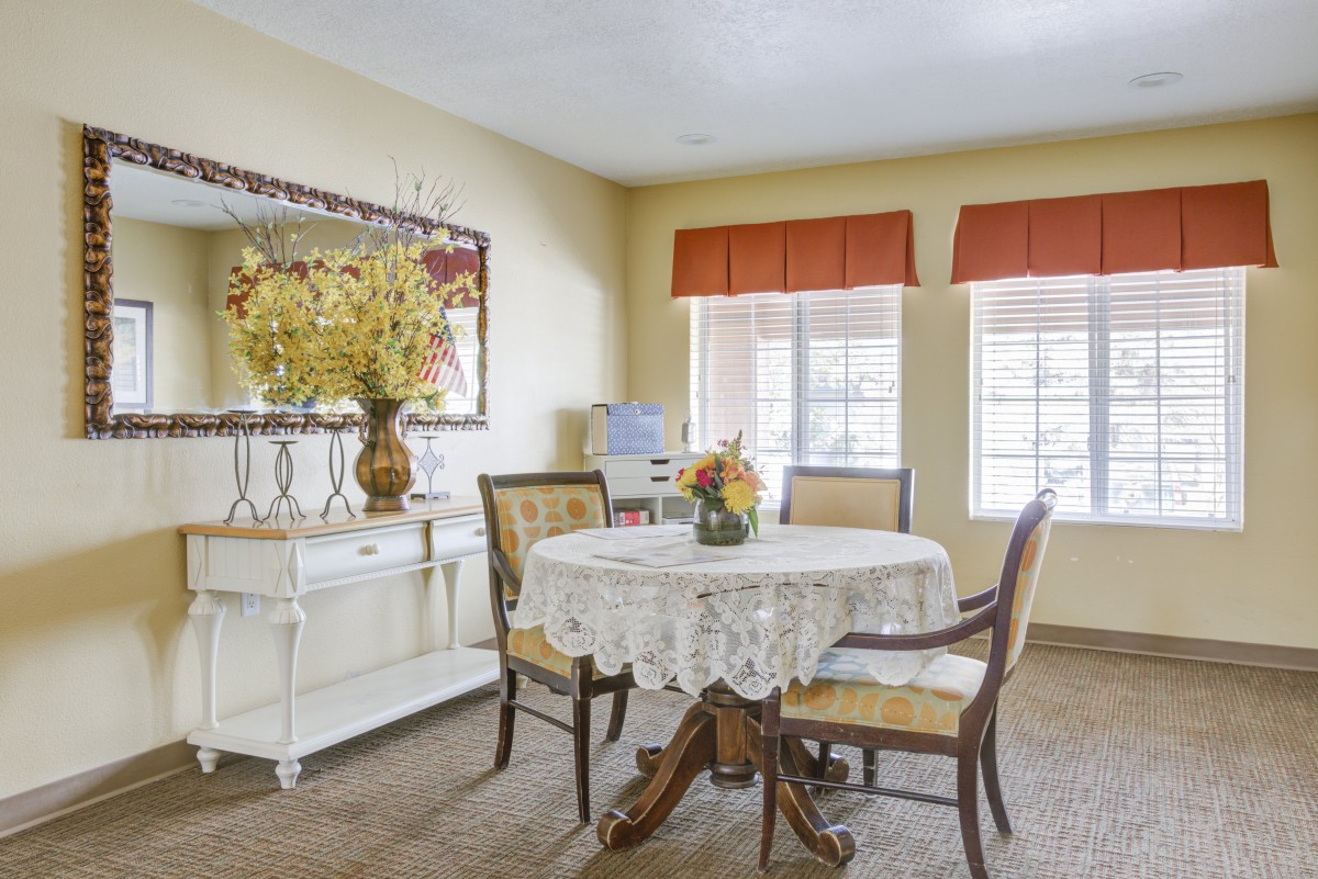A cozy dining room with a round table covered by a lace tablecloth and surrounded by four chairs, featuring a decorative mirror and floral arrangement.
