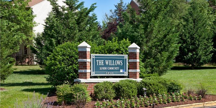 Entrance sign for The Willows senior community surrounded by greenery and brick pillars.