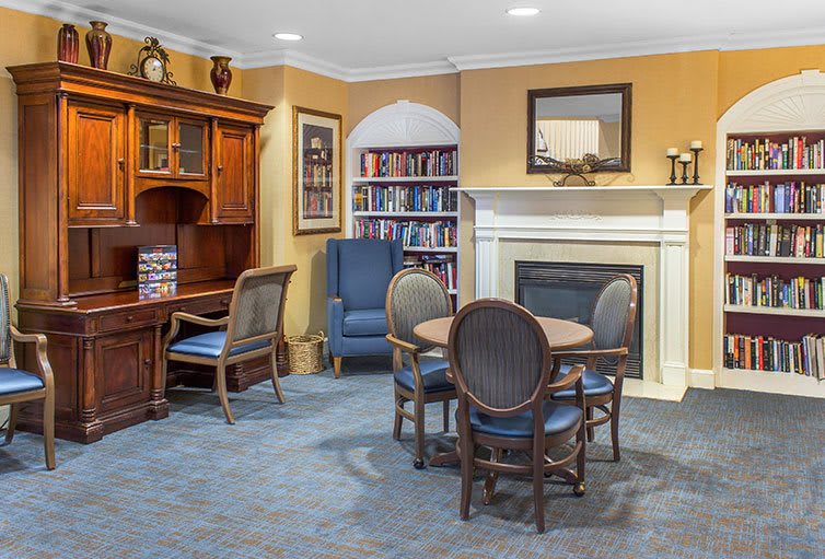 A cozy living room with a library corner, featuring a fireplace, bookshelf, wooden desk, and seating area.